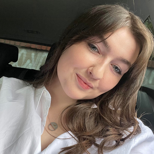 A headshot of a young woman with long brown hair, dressed in a white shirt, smiling with her head tilted towards the camera.