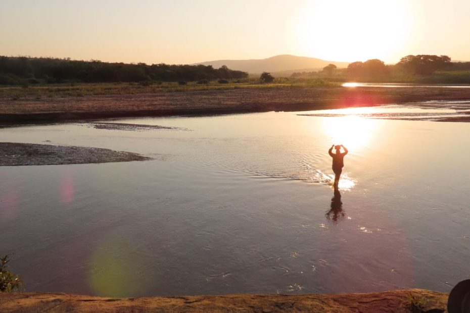 Photo of a human figure crossing a river at sunset or sunrise, in a wide open landscape