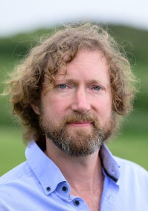 Headshot of a middle-aged man with wavy brown hair down to his ears and a beard, against a blurred background of grassy hills