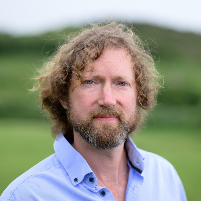 Headshot of a middle-aged man with wavy brown hair down to his ears and a beard, against a blurred background of grassy hills