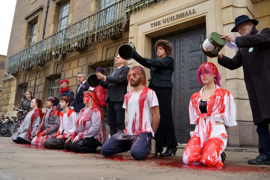 Photo of a row of people kneeling while another row behind them pour red liquid over their heads.