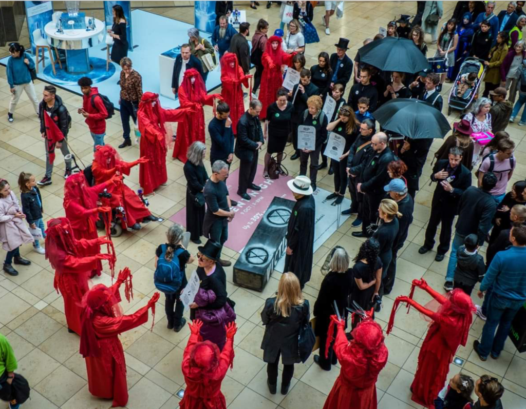 A crowd gathered as if in mourning stand around a casket painted with the Extinction Rebellion logo. And outer circle of figures are dressed in flowing red costumes.