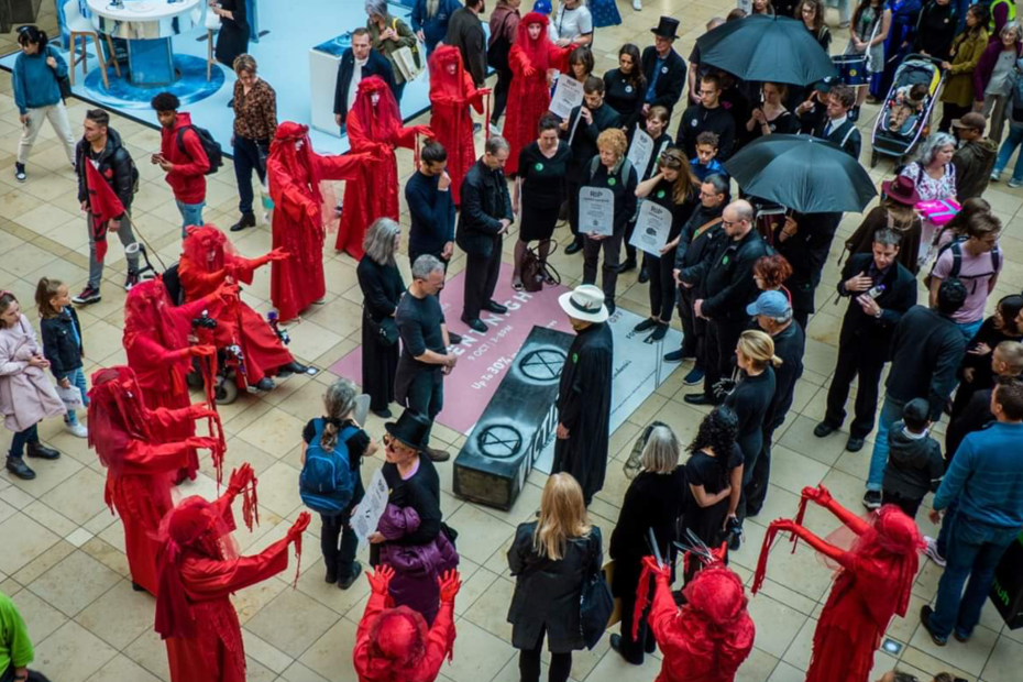 A crowd gathered as if in mourning stand around a casket painted with the Extinction Rebellion logo. And outer circle of figures are dressed in flowing red costumes.